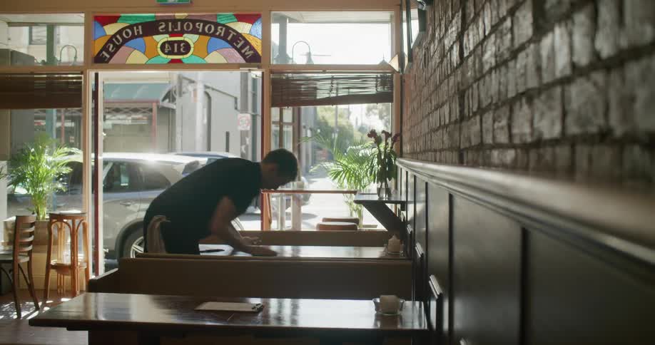 A man sprays and wipes down a restaurant booth located in a busy city as traffic passes by outside
