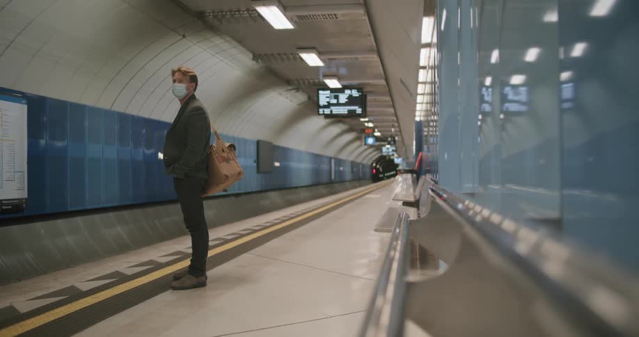 A man wearing a suit and face mask stands on a train platform waiting