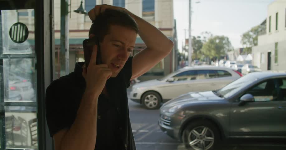 White man talking on the cellphone inside a building next to the door with people walking by and cars on the street