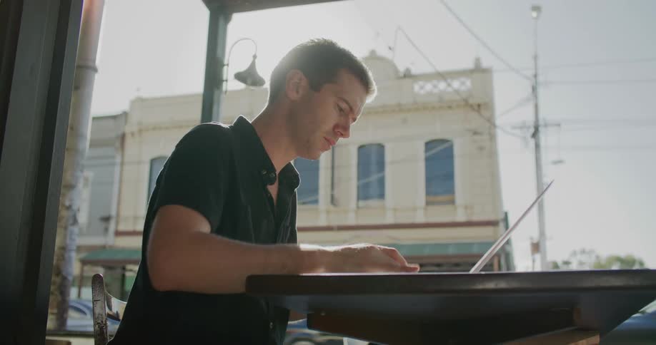 A man sits at a table in a restaurant and types on his laptop