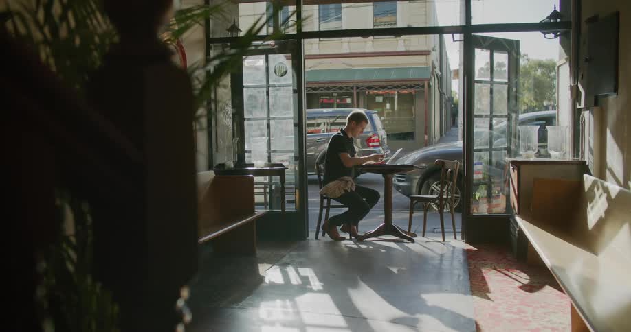 A man sitting at a table typing on a laptop then talking on a smart phone