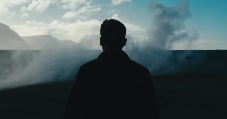 A man waves through a barren flatland towards a smoking vehicle