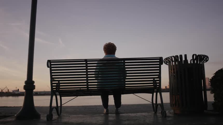 An older woman sits alone on a riverside bench at dusk