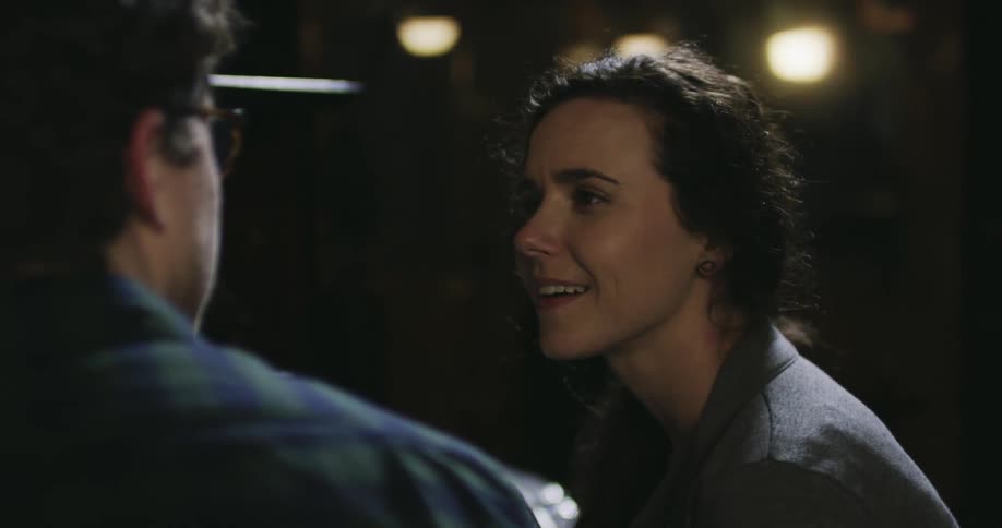 A man and woman talk in a dim room