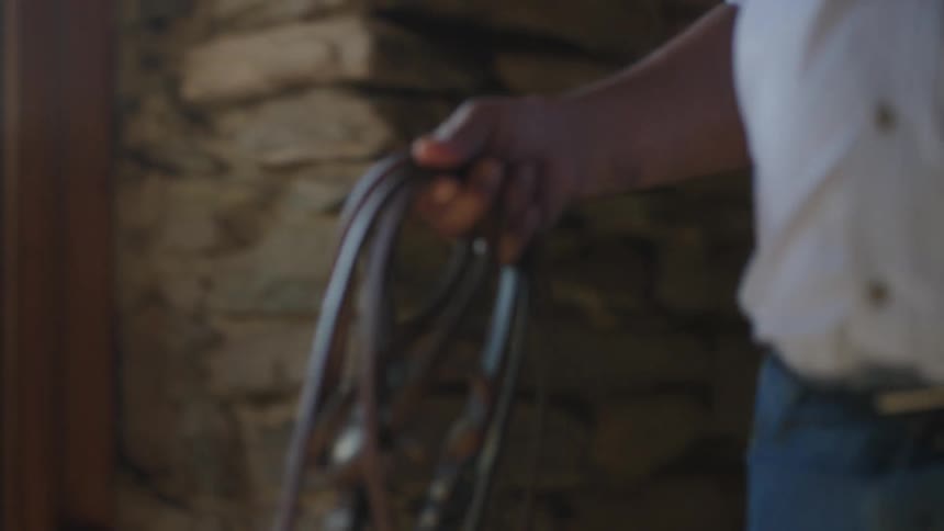 A cowboy wearing a cowboy hat walking in cobblestone stable with reins in his hand