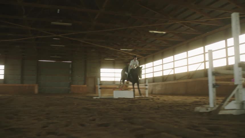 Person rides horse over jump in interior corral as another person watches