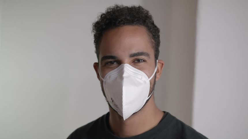 Young man puts on a white face mask while standing in a white room