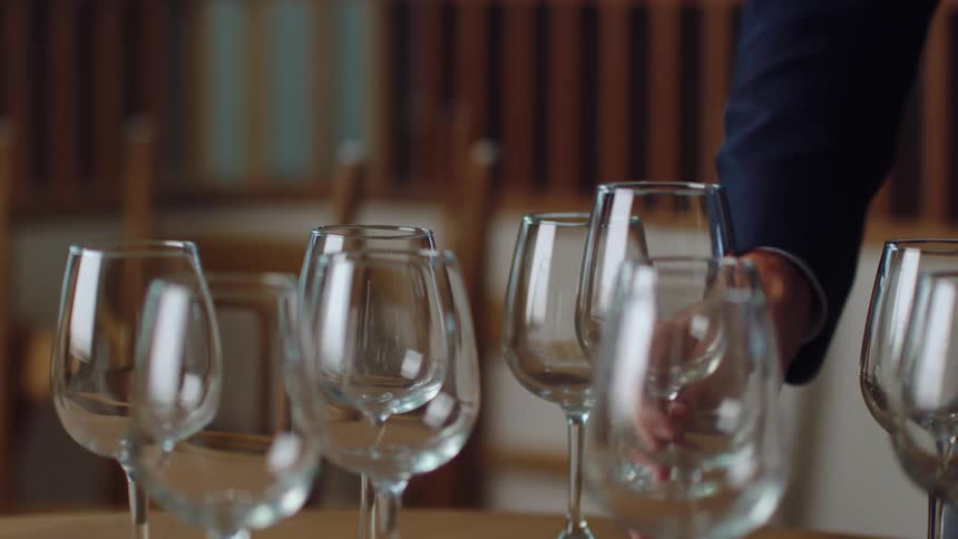 A person in a room placing a wine glass on a table with other wine glasses with chairs in the background