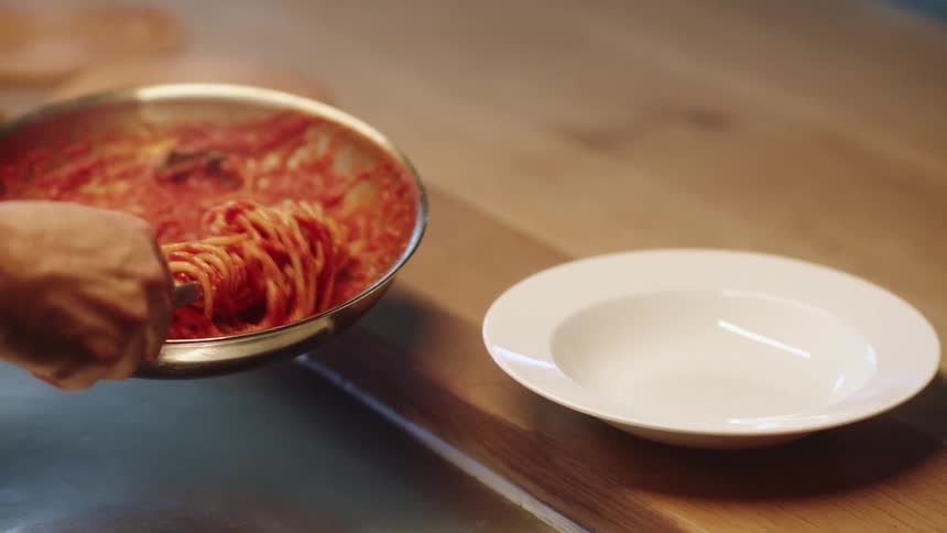 A person twirling spaghetti in a pan next to a table and a plate