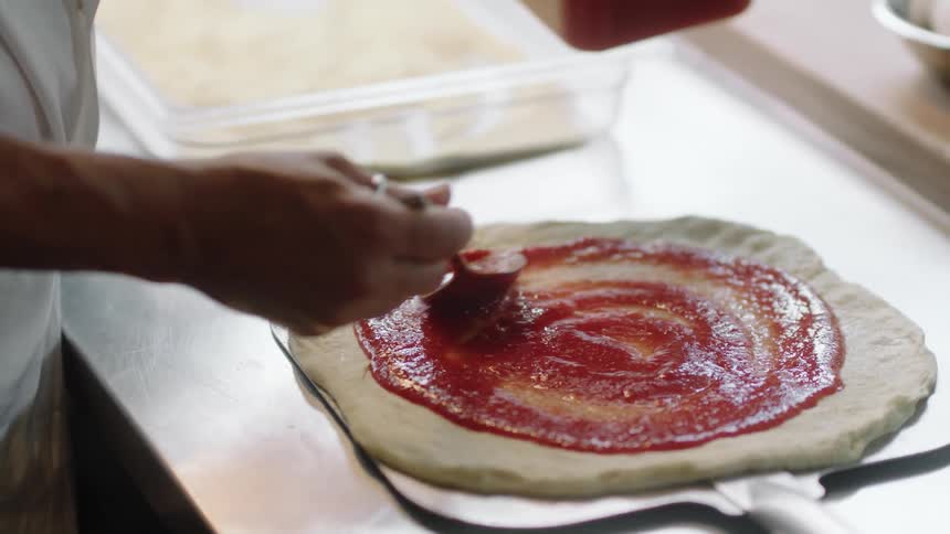 A chef spoons tomato sauce onto a pizza in a restaurant kitchen