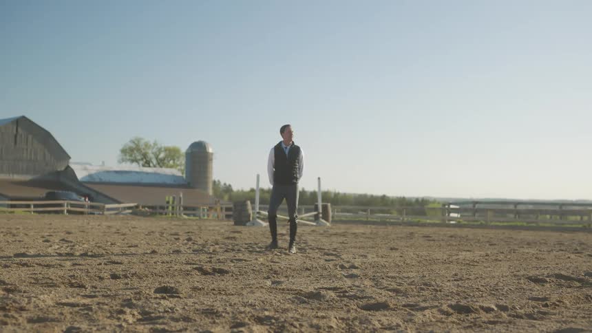 A man stands in the middle of a riding arena and watches as a young woman rides a horse