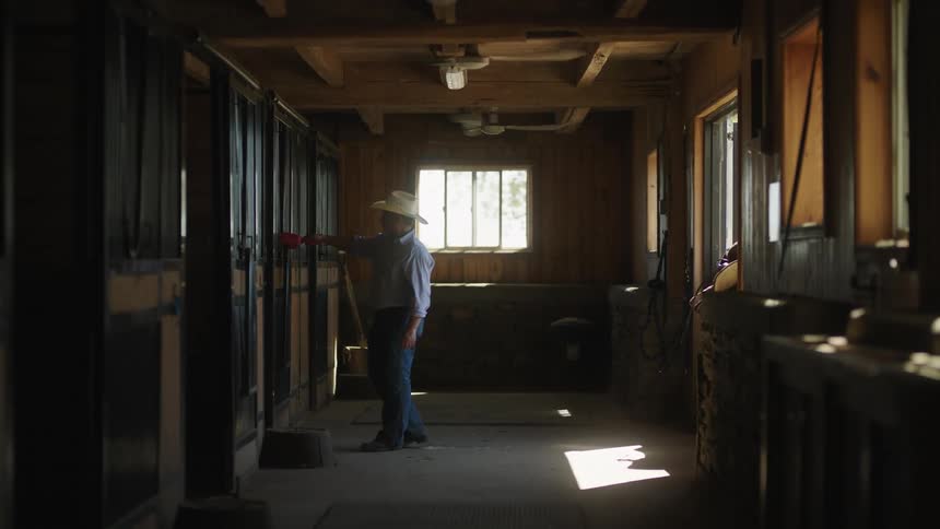 A man wearing a cowboy hat and jeans stands outside a stall in a stable