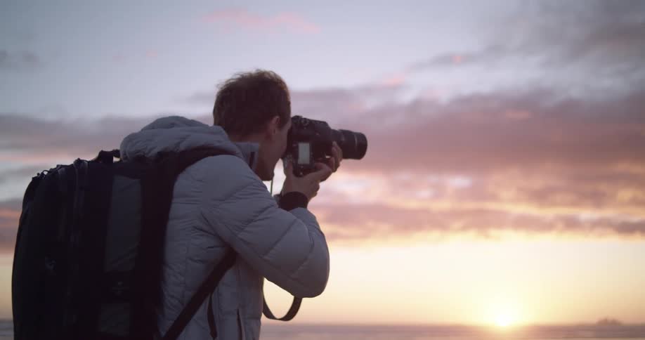 A man holds a large professional camera to his face looking at an ocean sunset horizon