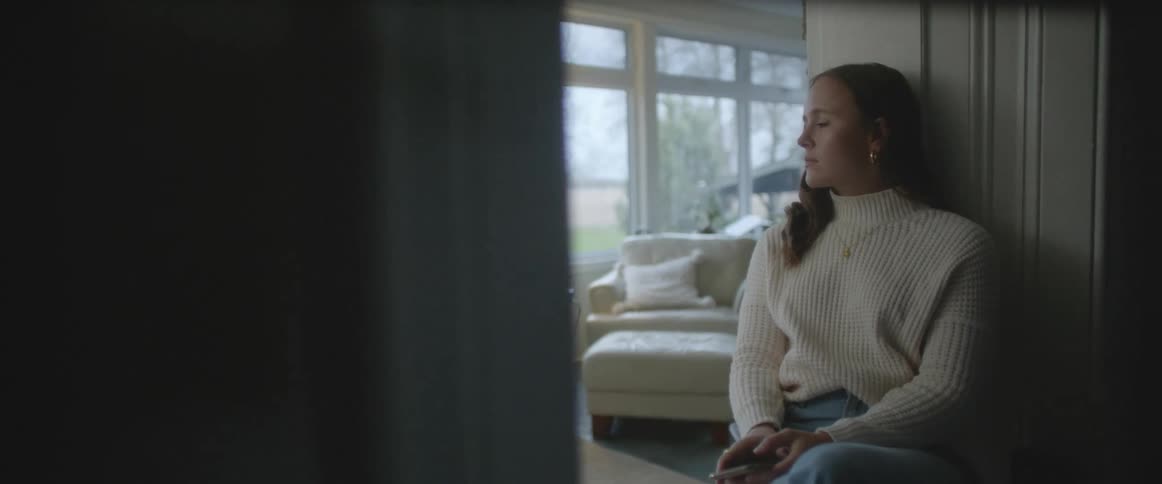 A woman with a sweater sitting in a doorway in a room with windows and a lamp in the background