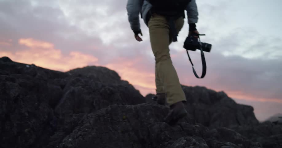 Man walking over rocks towards red sky while holding a professional camera