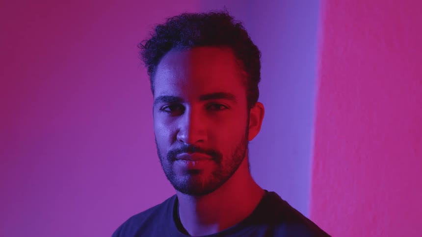 A bearded man stands looking straight ahead opening and closing his eyes as pink lights flash