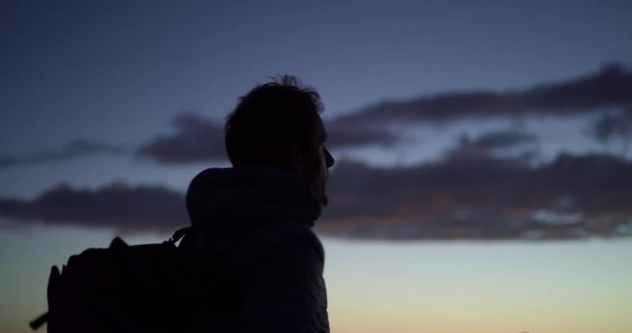 Man partly silhouetted against red sky puts on backpack