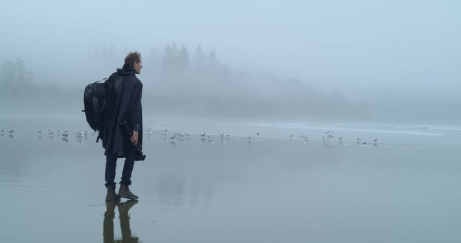 A man walks across a beach with seagulls carrying a camera as the rain falls