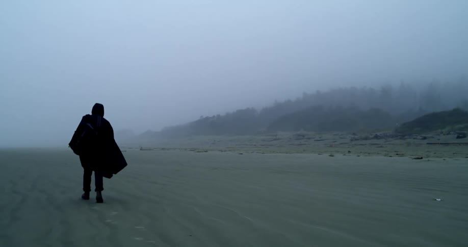 A man in rain coat is hiking along beach in thick ocean fog
