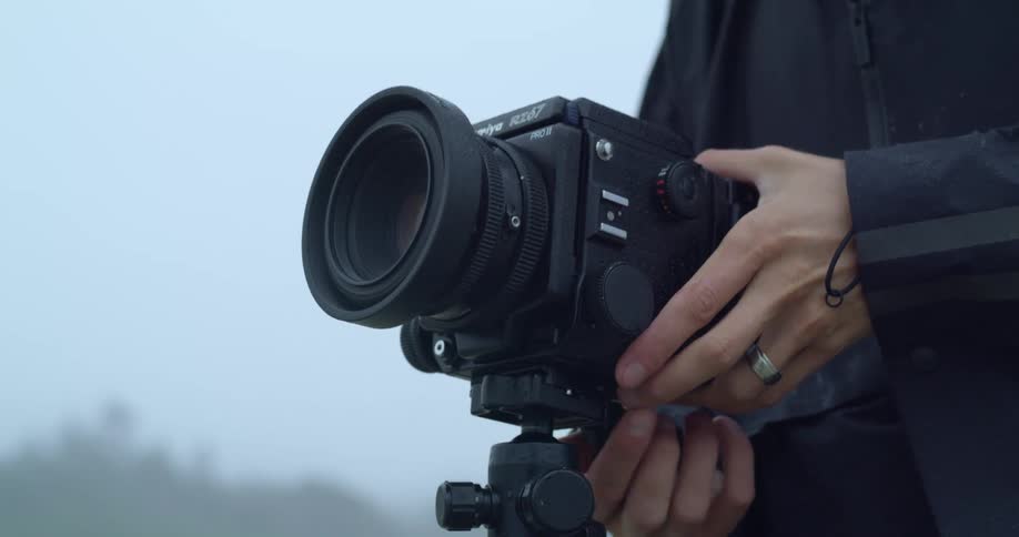 A photographer in a raincoat unscrews a camera from a tripod standing outdoors on a rainy day
