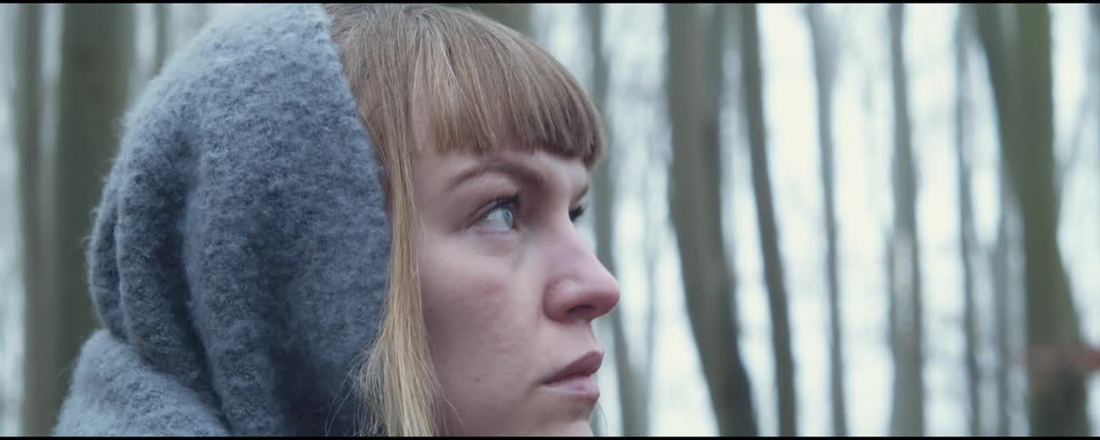 A woman wears a gray scarf over her head as she stands in a forest filled with tall barren trees