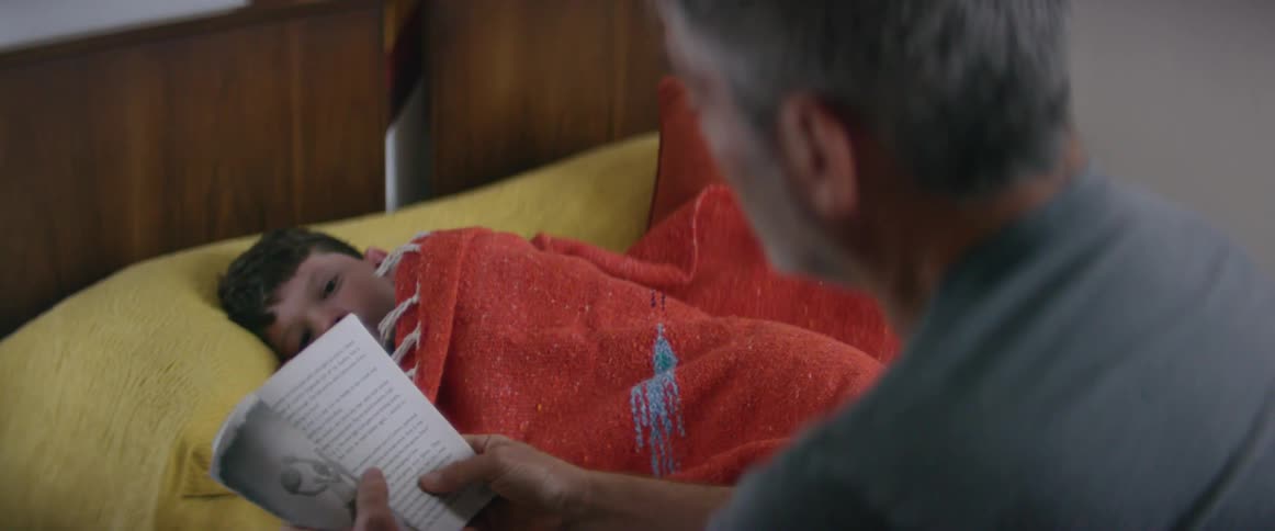 A little boy lies in bed as his dad sits on the bed and reads him a story