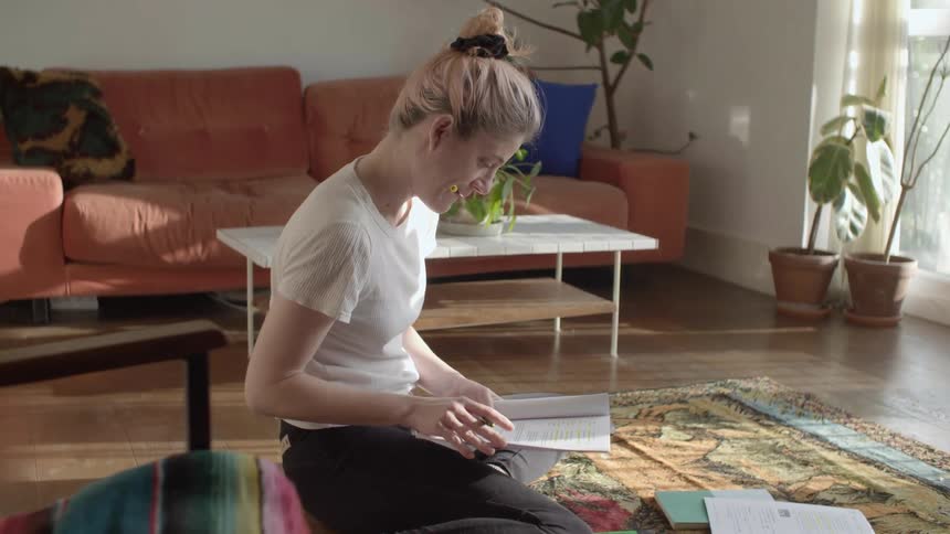 A woman sits on a colorful rug on her apartment floor with homework spread in front of her