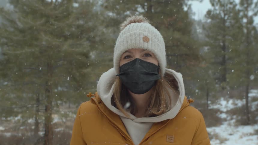 A woman wears a yellow parka and black face mask she she walks through a forest as snow falls