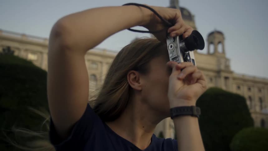 A woman adjusting her camera settings and taking a photo with an architectural building behind her