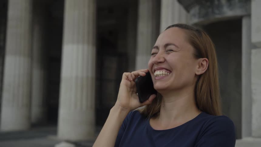 A woman standing and talking on a smart phone with a building with pillars in the background