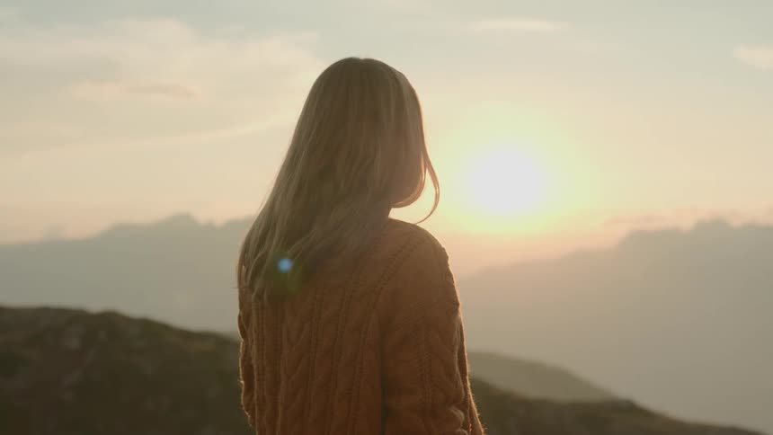 Woman stands looking toward sun shining over low mountains in red tinted sky