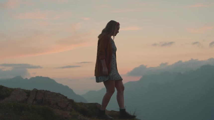 A woman with a dress walking on a rocky terrain with a mountain range the sky and clouds in the background
