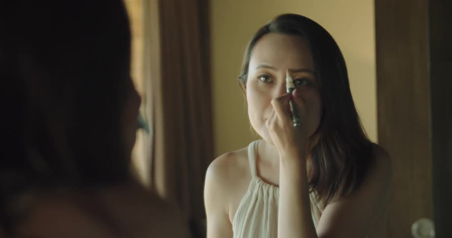 Woman with white sleeveless top looks in mirror and puts on makeup