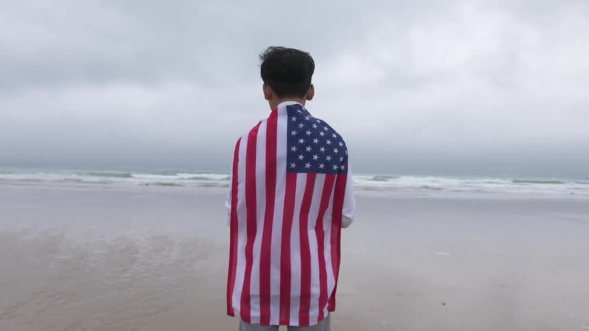 A man with an american flag draped around his shoulders looks out contemplatively at the incoming waves while standing on a beach on a cloudy day