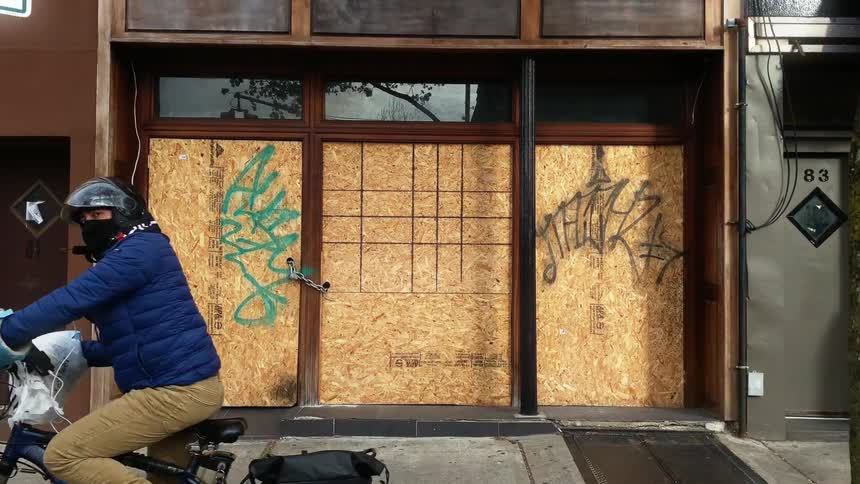 Man riding a bicicyle passes in front of the boarded windows and door of a street store