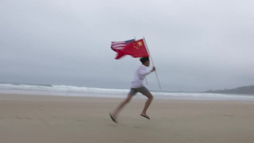 You male wearing white shirt and shorts running across a beach carrying a chinese flag and an american flag