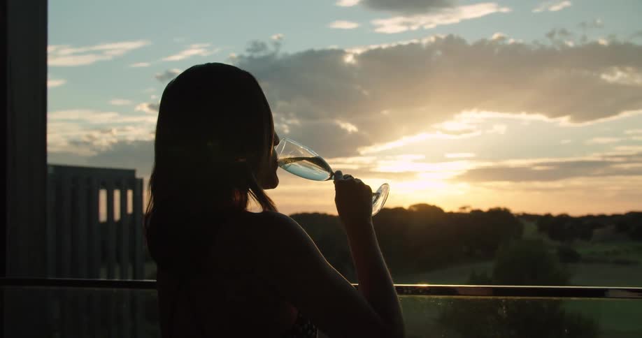 A woman on a balcony drinks wine from a wine glass as she gazes out over the countryside below in the evening