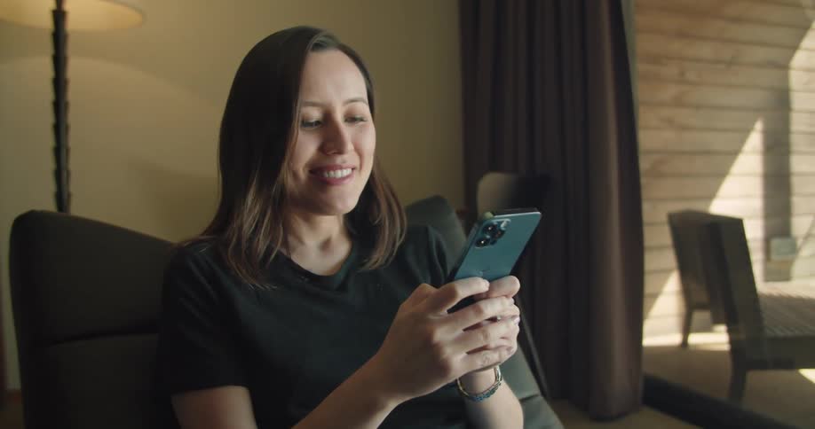 A woman sitting at home smiles as she uses her cell phone