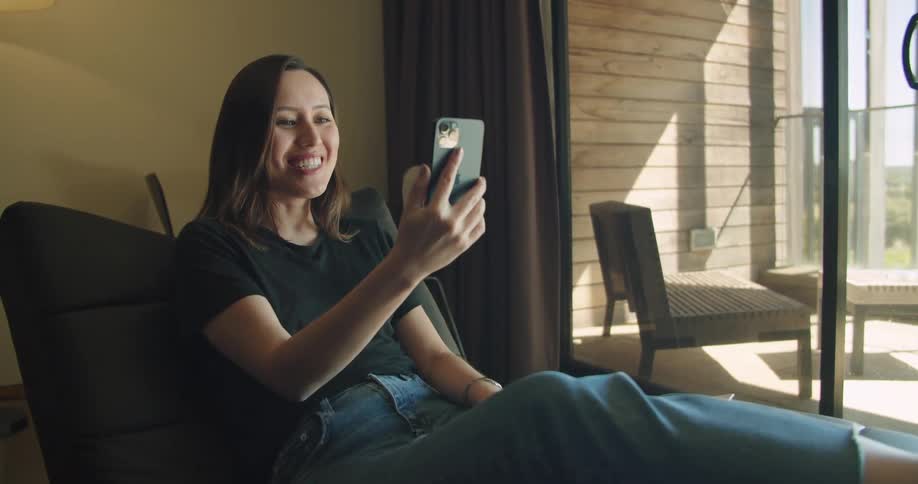 Woman with black t shirt and jeans sits in chair and makes a video call on smartphone in the living room at home