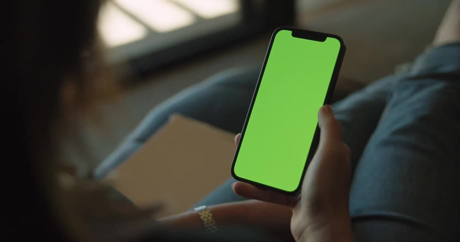 A woman scrolls through her smartphone while sitting at home by a large window