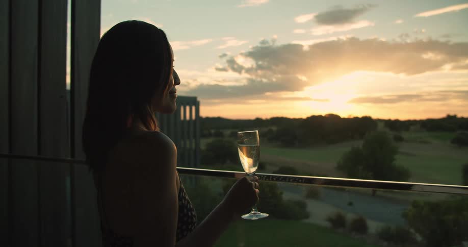A woman stands on a balcony holding a wine glass looking at the sunset