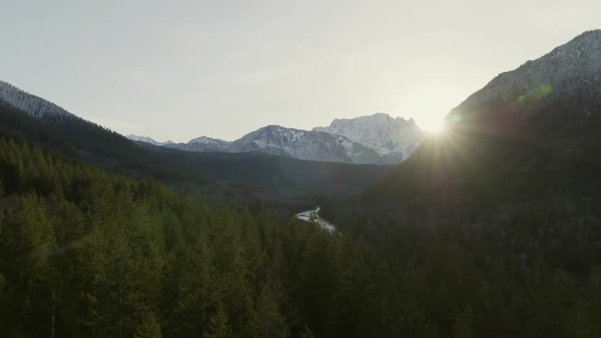 The sun peeks out from behind mountains to illuminate a winding river through a vast forest valley