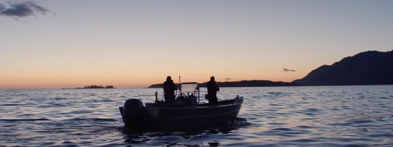 A silhouette of two people on a boat on a body of water with land the sky and clouds in the background
