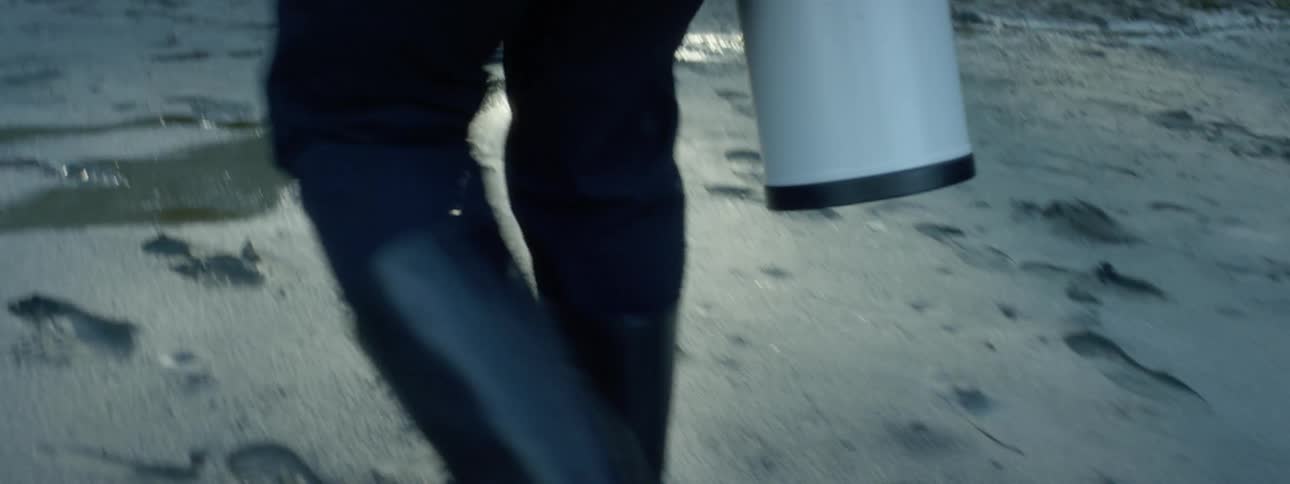 A photographer wearing boots walks on wet sand carrying a camera zoom lens