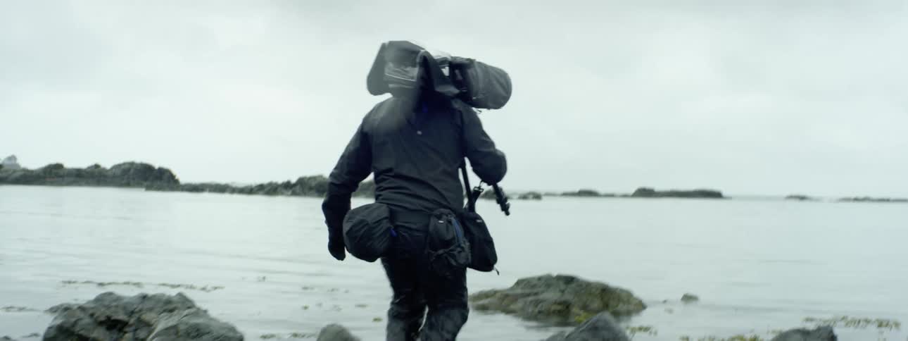 Cameraman carrying camera over his shoulder walks on the coast next the ocean cliffs