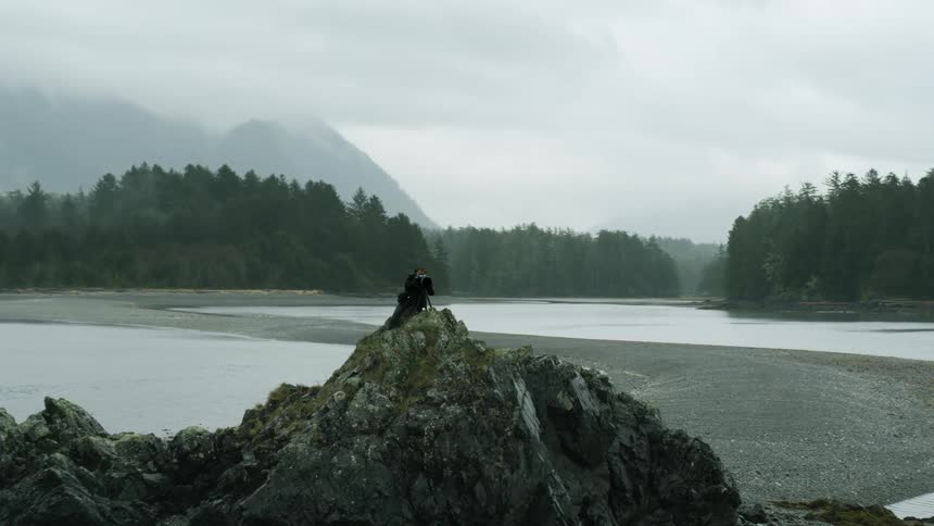 Male photographer points his camera while standing on a rock at a coastline
