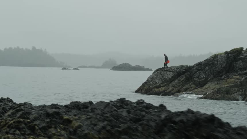 A man carries an orange item over a rocky terrain to the coastline