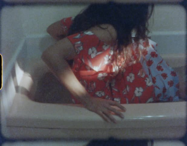 A woman wearing a dress steps in to a bathtub filled with water and lies down