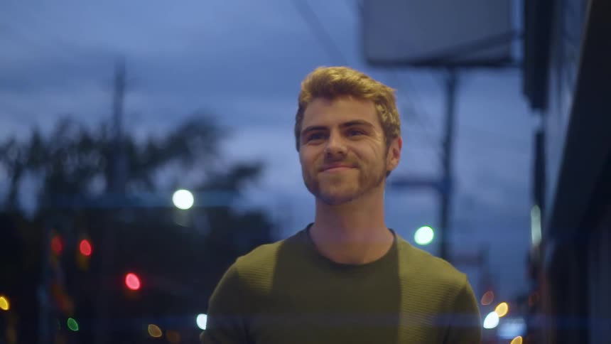 A man standing outside of a building in a city at night smiles as he gazes out of frame