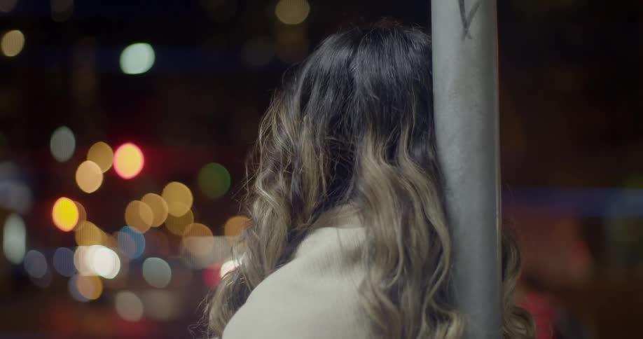 A woman with long curly hair leaning against a metal pole while pedestrians walk by in a city